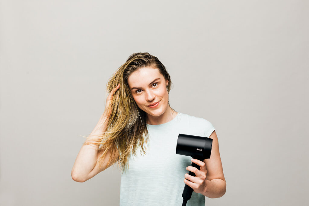 A woman with long blonde hair blow dries her hair with the shark style iq hairdryer protecting it from heat with ionised technology